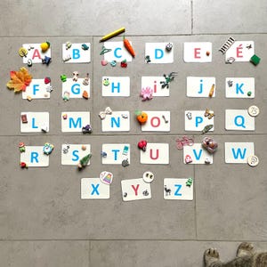 May include: Alphabet magnets with various decorative elements. Each letter is on a white rectangle, with colorful embellishments like a lemon, a leaf, a motorcycle, and a bicycle. The letters are in blue and red.