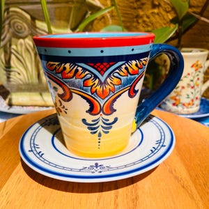 May include: A ceramic coffee mug and saucer set. The mug has a blue handle and is decorated with a colorful floral design in red, yellow, and blue. The saucer is white with a blue border. The set is on a wooden surface.