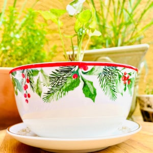 May include: A white ceramic bowl with a red rim and a matching saucer. The bowl is decorated with green holly leaves and red berries. The bowl is sitting on a wooden surface.