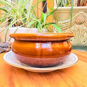 May include: A glossy, amber-colored ceramic bowl with a rounded shape and a wide, smooth rim. The bowl rests on a white saucer featuring a delicate, intricate gold-toned filigree pattern along its edge, set against a warm wooden surface.
