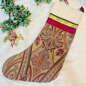 May include: A Christmas stocking with a paisley pattern in shades of red, gold, and brown. The top of the stocking is white with a red and gold ribbon detail. A festive holiday decoration.