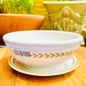 May include: A white ceramic bowl with a matching saucer. The bowl has a blue rim and is decorated with a gold geometric pattern and leaf design. The saucer has a gold rim and a matching leaf design.