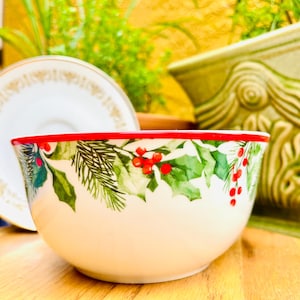 May include: A cream-colored ceramic bowl with a red rim, decorated with green holly leaves, red berries, and pine needles. The bowl is on a wooden surface, with a white plate and green plant pots in the background.