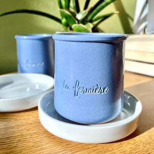 May include: Two light blue ceramic jars with the text "la fermiere" embossed on the side. The jars are sitting on white ceramic saucers. A third jar is visible in the background. The jars are on a wooden surface.