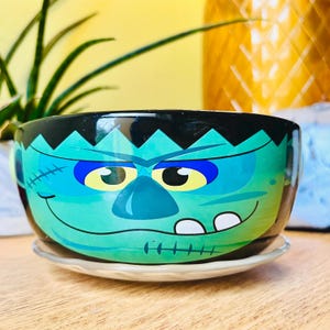 May include: A round ceramic bowl featuring a green monster face with a black top, blue eye shadows, yellow eyes, and stitched scars. The bowl has a wide mouth, a curved smile with two visible white teeth, and sits on a small, clear glass saucer.