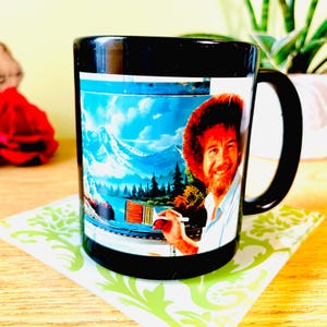 May include: Black ceramic coffee mug featuring a colorful image of a smiling man with an afro and a landscape painting. The mug has a black handle and rim. The image includes mountains, trees, and a lake. The mug is on a green and white patterned coaster.