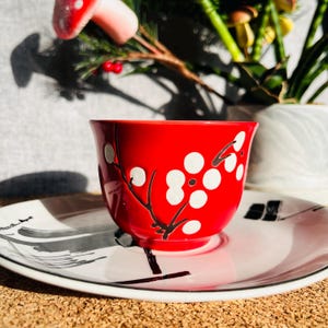 May include: A red teacup with a white floral design. The teacup is sitting on a white saucer with a black design.