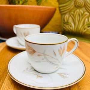 May include: White teacup and saucer set with gold trim and wheat-like design. The teacup has a handle and sits on a matching saucer. A second teacup and saucer are visible in the background. The set is on a wooden surface.