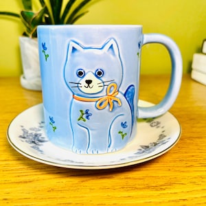 May include: A light blue ceramic mug featuring a raised design of a cat with a yellow bow and orange collar. The mug sits on a matching saucer with a floral pattern. The mug has a curved handle and is decorated with small blue flowers.