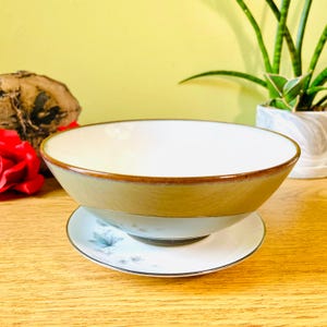 May include: A ceramic bowl with a light beige exterior and a white interior, resting on a matching saucer. The bowl has a brown rim and sits on a white saucer with a blue floral design. The bowl and saucer are on a wooden surface.