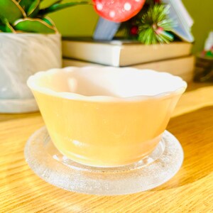 May include: A yellow-orange bowl with a scalloped rim sits on a clear glass coaster. The bowl has a smooth exterior and a white interior. The coaster is round and has a textured, frosted appearance. The items are on a wooden surface.