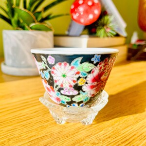 May include: A small, black ceramic cup with a floral design in pink, red, blue, and yellow. The cup sits on a clear, glass pedestal. The cup has a white rim and is likely used for tea or other beverages.