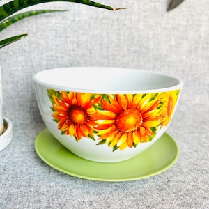 May include: White ceramic bowl with a green saucer. The bowl has a sunflower design with yellow and orange petals.