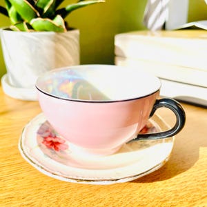 May include: A pink teacup and saucer set. The teacup has a black handle and rim, and the saucer features a floral design. The teacup and saucer are sitting on a wooden surface. A plant in a white pot is in the background.