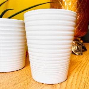 May include: Two white cylindrical plant pots with a horizontal ribbed texture. The pots have a matte finish and a tapered shape, narrowing slightly toward the base. The surface of each pot features consistent, evenly spaced ridges.