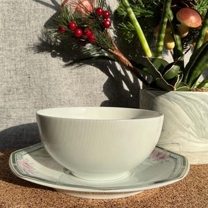 May include: White ceramic bowl with a green and floral patterned saucer. The bowl and saucer are sitting on a brown surface.