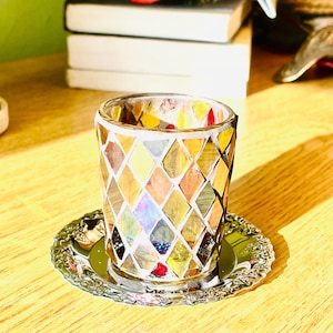 May include: A decorative glass votive candle holder with a mosaic design in shades of gold, amber, and red. The candle holder sits on a silver-colored, ornate, round coaster. The mosaic pattern is made of small, irregularly shaped pieces of glass.
