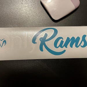May include: A white sticker with the word "Rams" in teal script. The sticker also features a teal ram's head logo and the word "only" in a faded font. The sticker is rectangular and is on a black surface.