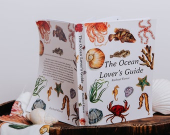 May include: An open book titled "The Ocean Lover's Guide" with colorful illustrations of marine life. The cover features a large seashell, crab, seahorse, and starfish. The book is on a wooden tray with a seashell and a cloth.