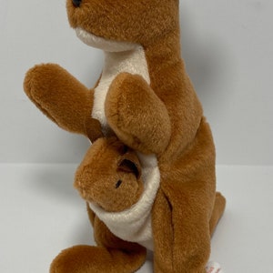 May include: A brown and white kangaroo beanie baby with a joey in its pouch. The beanie baby has a red and white Ty tag attached to its ear that reads "Beanie Original Baby".