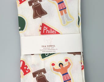 May include: White tea towel with a repeating pattern of the Liberty Bell, a boxer, and Phillies pennants. The tea towel is wrapped with a white band that reads "TEA TOWEL" and "SPOGAS ART".