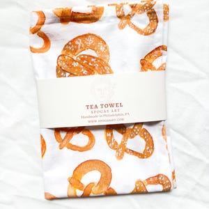 May include: White tea towel with a repeating pattern of brown pretzel illustrations. The text "TEA TOWEL SPOGAS ART Handmade in Philadelphia, PA www.spogasart.com" is printed on a white label attached to the towel.