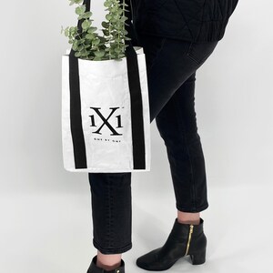 May include: A white tote bag with black straps and the logo "1x1 ONE BY ONE" printed on the front. The bag is being held by a person wearing black jeans and a black jacket.