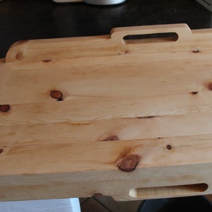 May include: A large, rectangular wooden chopping board with a handle. The board is made of light wood with a natural finish and has several knots. The board is ideal for preparing food or serving snacks.