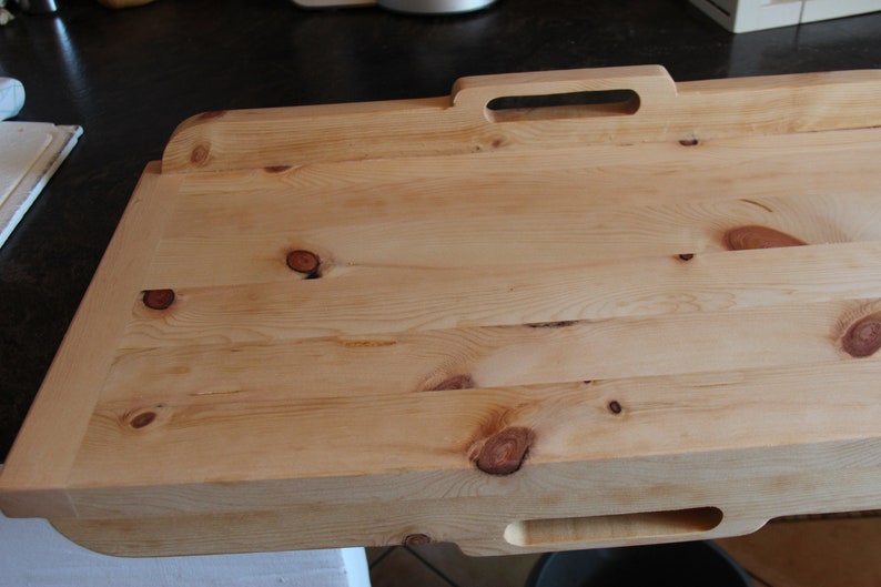 May include: A large, rectangular wooden chopping board with a handle. The board is made of light wood with a natural finish and has several knots. The board is ideal for preparing food or serving snacks.