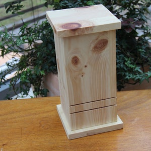 Puede incluir: Una caja de madera clara con una base cuadrada y una tapa rectangular. La caja tiene un acabado de madera natural y está hecha de madera maciza.