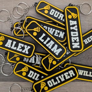 May include: A selection of black and yellow lacrosse keyrings, each with a name in white lettering. The keyrings feature a lacrosse stick and ball design. The keyrings are rectangular with rounded edges and attached to silver keyrings.