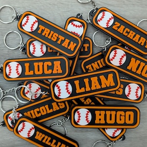 Puede incluir: Una colección de llaveros personalizados con temática de béisbol. Cada llavero presenta un gráfico de béisbol y un nombre en letras naranjas sobre un fondo negro. Los llaveros están sujetos a llaveros plateados.