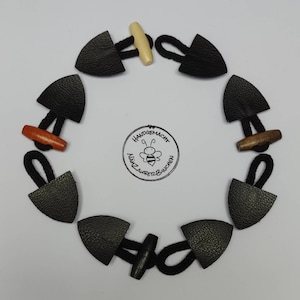 Könnte beinhalten: Ein Satz von acht schwarzen Leder-Toggle-Knöpfen mit Holz-Toggle. Die Toggle sind in verschiedenen Farben, darunter Braun, Orange und Weiß. Die Knöpfe sind in einem Kreis auf einer weißen Oberfläche angeordnet. Der Text "Handgemacht Nähzauberbinchen" ist in der Mitte des Kreises sichtbar.