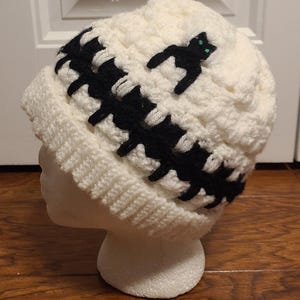 May include: White crocheted beanie with a black crocheted band and a black cat applique with green eyes.