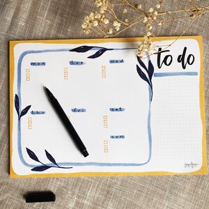 Puede incluir: Un planificador semanal blanco con un borde amarillo y acentos florales azules en acuarela. El planificador tiene una sección de lista de tareas pendientes y una sección diaria para cada día de la semana. El planificador está sobre una superficie de tela marrón.