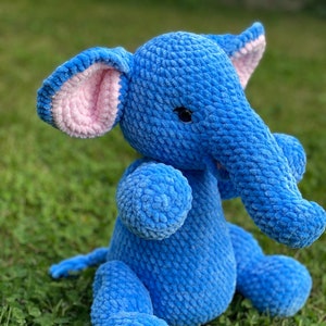 May include: A blue crocheted elephant plush toy with pink ears. The elephant is sitting on a bed of green grass.