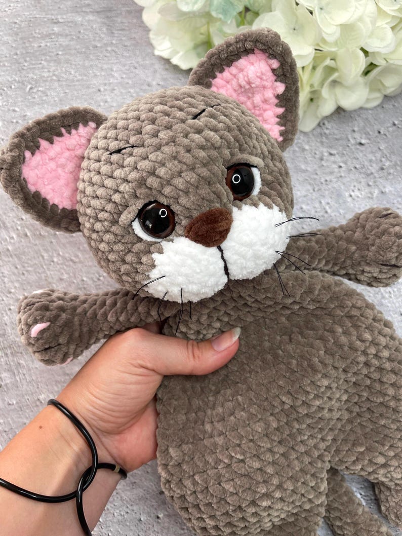 May include: A handmade plush cat toy in shades of brown, with pink inner ears and white accents on the muzzle. The cat has large, expressive eyes and black whiskers. The toy is soft and cuddly, perfect for children.