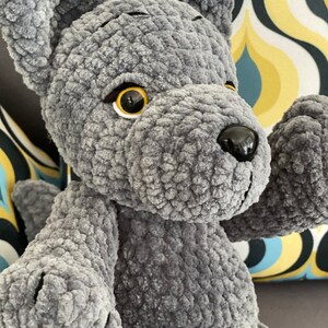 Pu&ograve; includere: Un giocattolo di peluche di lupo grigio all'uncinetto con occhi neri e naso nero. Il lupo &egrave; seduto su un divano con un tessuto a motivi.