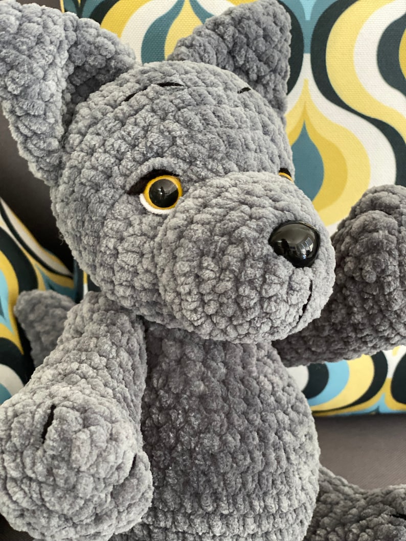 Pu&ograve; includere: Un giocattolo di peluche di lupo grigio all'uncinetto con occhi neri e naso nero. Il lupo &egrave; seduto su un divano con un tessuto a motivi.