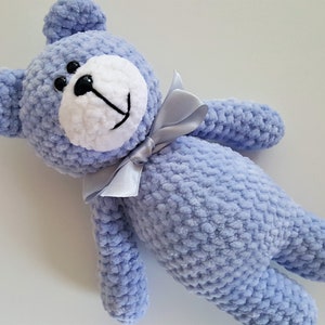 Puede incluir: Un oso de peluche azul tejido a crochet con una cara blanca y ojos negros. El oso lleva un lazo de sat&eacute;n azul claro.