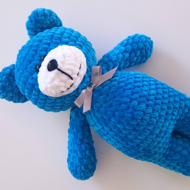 Puede incluir: Un oso de peluche azul tejido a crochet con una cara blanca y ojos negros. El oso tiene una cinta gris atada alrededor de su cuello.
