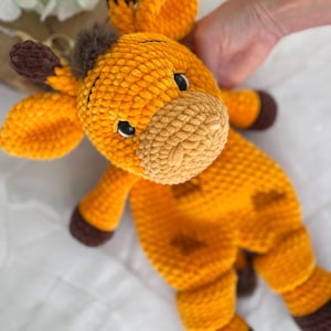 以下が含まれることがあります： 黄色と茶色の手作りのぬいぐるみキリン。キリンは、ダークブラウンの蹄、耳、斑点を持つ、かぎ針編みのテクスチャの外観をしています。おもちゃの目は黒で、鼻は明るいタン色です。