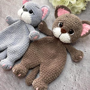May include: Two handmade plush cat toys. One is gray with pink and white accents, the other is brown with pink and white accents. Both have large eyes, small paws, and are lying down. The toys are made of a soft, textured material.