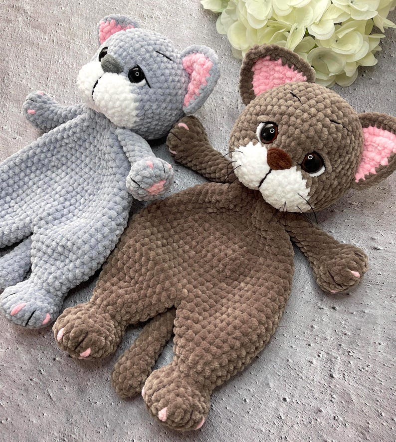 May include: Two handmade plush cat toys. One is gray with pink and white accents, the other is brown with pink and white accents. Both have large eyes, small paws, and are lying down. The toys are made of a soft, textured material.
