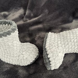 May include: A pair of gray and white crocheted slipper boots. The boots have a fluffy texture and a gray trim around the top.