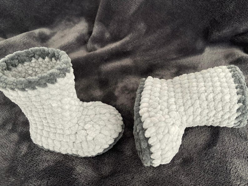 May include: A pair of gray and white crocheted slipper boots. The boots have a fluffy texture and a gray trim around the top.