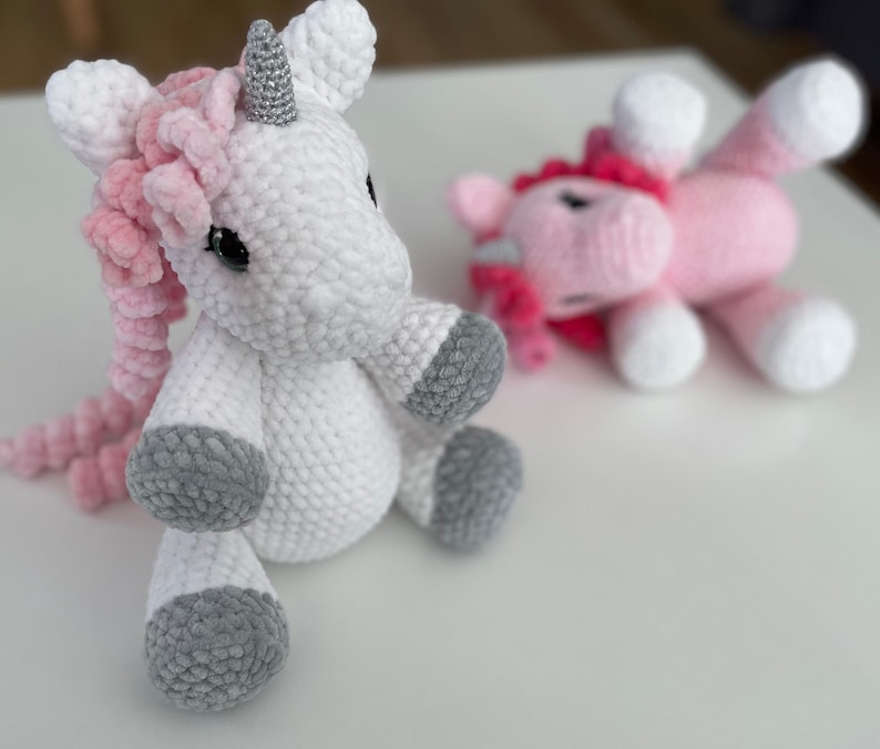 May include: Two crocheted unicorn toys. One is white with a pink mane and tail, the other is pink with a pink mane. Both have grey hooves.