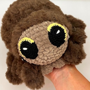 May include: A brown crocheted spider plush toy with large black eyes and yellow accents. The spider has a fuzzy texture and is being held by a hand.