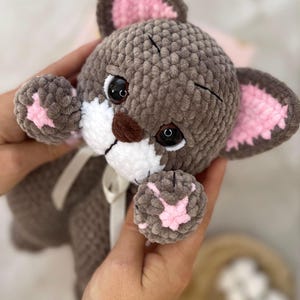May include: A handmade plush cat toy in shades of gray, white, and pink. The cat has large black eyes, a white muzzle, and pink accents on its ears and paws. A white ribbon is tied around its neck.