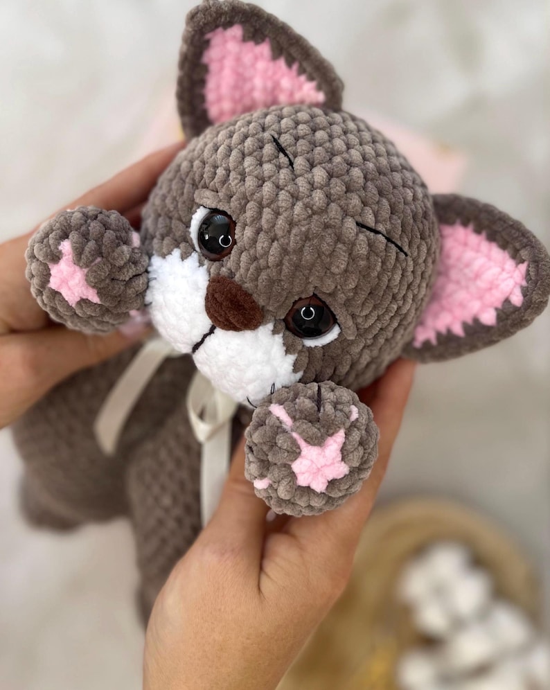 May include: A handmade plush cat toy in shades of gray, white, and pink. The cat has large black eyes, a white muzzle, and pink accents on its ears and paws. A white ribbon is tied around its neck.